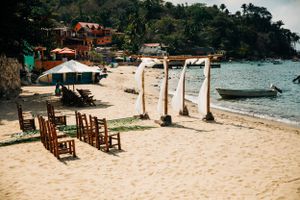 Wooden rocking chairs and draped fabric posts line a tropical beach with clear waters and hillside villas in the background.