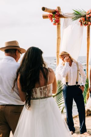 A beach wedding ceremony under a bamboo arch decorated with coral flowers and white fabric, overlooking the ocean.