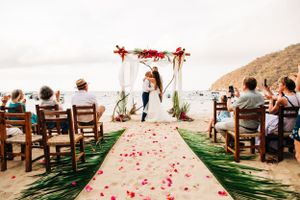 Beach wedding ceremony with wooden chairs and rose petals scattered on a green aisle runner leading to floral arch.