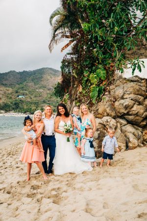 A group celebrating at a tropical beach wedding with palm trees and rocky cliffs in the background.