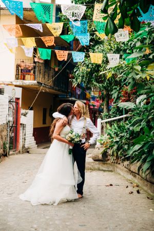 A wedding couple embraces under colorful Mexican papel picado banners hanging on a charming street with greenery.