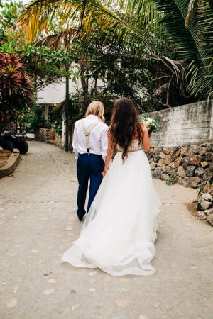 A wedding couple walks down a tropical palm-lined path, with the bride's flowing white dress trailing behind them.
