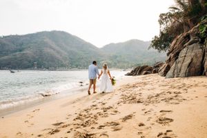 A couple walks hand in hand with their dog along a scenic beach with mountains and rocky cliffs in the background.