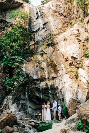 A scenic wedding ceremony takes place in front of a majestic waterfall cascading down rocky cliffs surrounded by lush greenery.