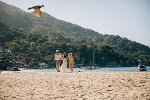 A seagull flies over beachgoers walking along a sandy shore with boats and mountains in the background.