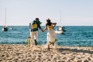 Couple in casual wedding attire runs joyfully along beach with boats visible in turquoise ocean background.