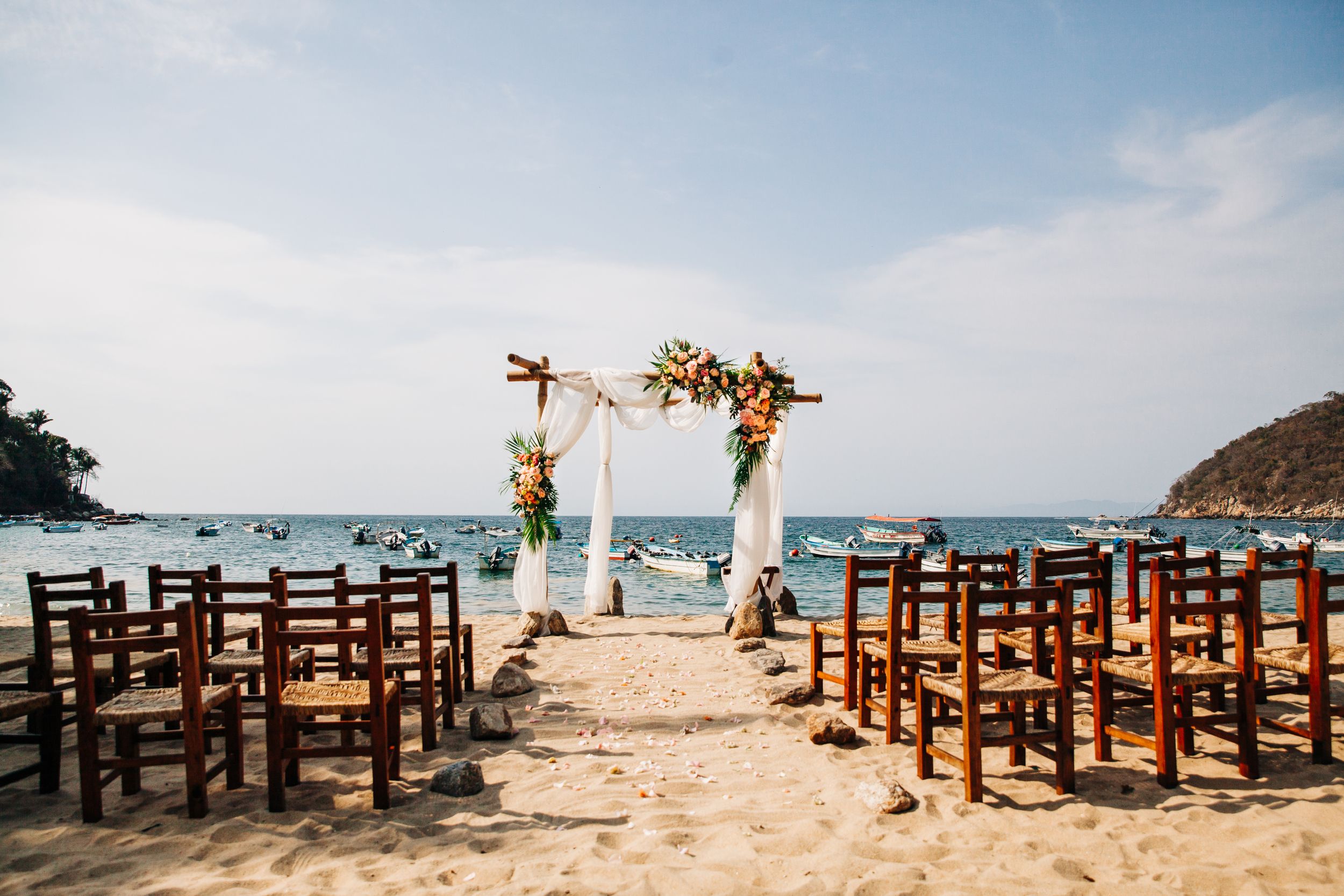 Beautiful beach wedding setup with wooden chairs and floral arch overlooking the ocean on a sunny day.
