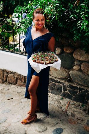 A person in a navy blue dress holds a tray of appetizers while standing on a stone path near a garden wall.