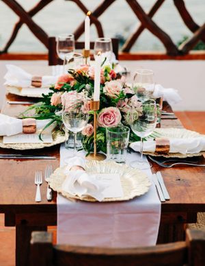 Elegant wedding table setting with pink roses centerpiece, gold candle holder, and blush runner on rustic wooden table.