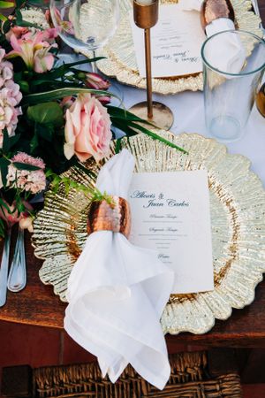 Elegant wedding place setting with gold charger plate, pink roses, and white menu card on wooden table.