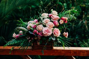 A stunning floral arrangement of pink and peach roses with tropical ferns on a wooden surface against greenery.