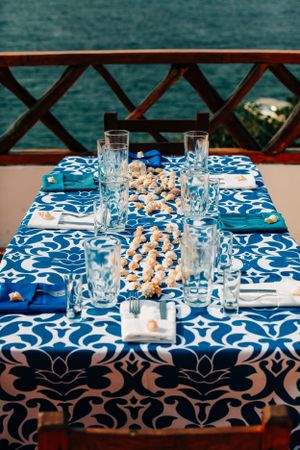A blue and white patterned tablecloth with crystal glasses and seashells set for a seaside wedding reception.