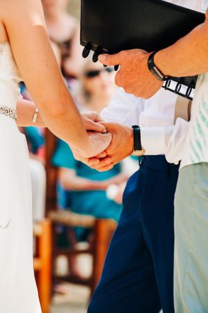 A close-up view of joined hands during a wedding ceremony, with white dress and navy suit visible in the frame.