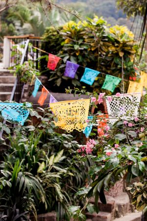 Colorful papel picado banners hang among lush tropical plants and foliage in a garden setting.