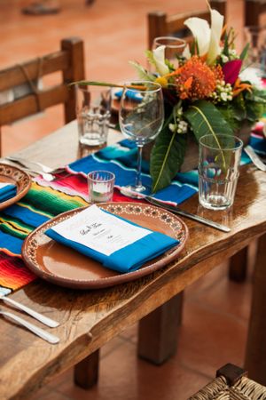 Mexican themed table setting with colorful serape runner, blue napkins and floral centerpiece on rustic wooden table.