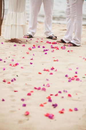 Pink and purple flower petals scattered on sand with feet wearing white pants and sandals at a beach wedding ceremony.