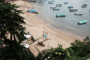 Small boats anchored in turquoise water near a sandy beach with palm trees and umbrellas on shore.