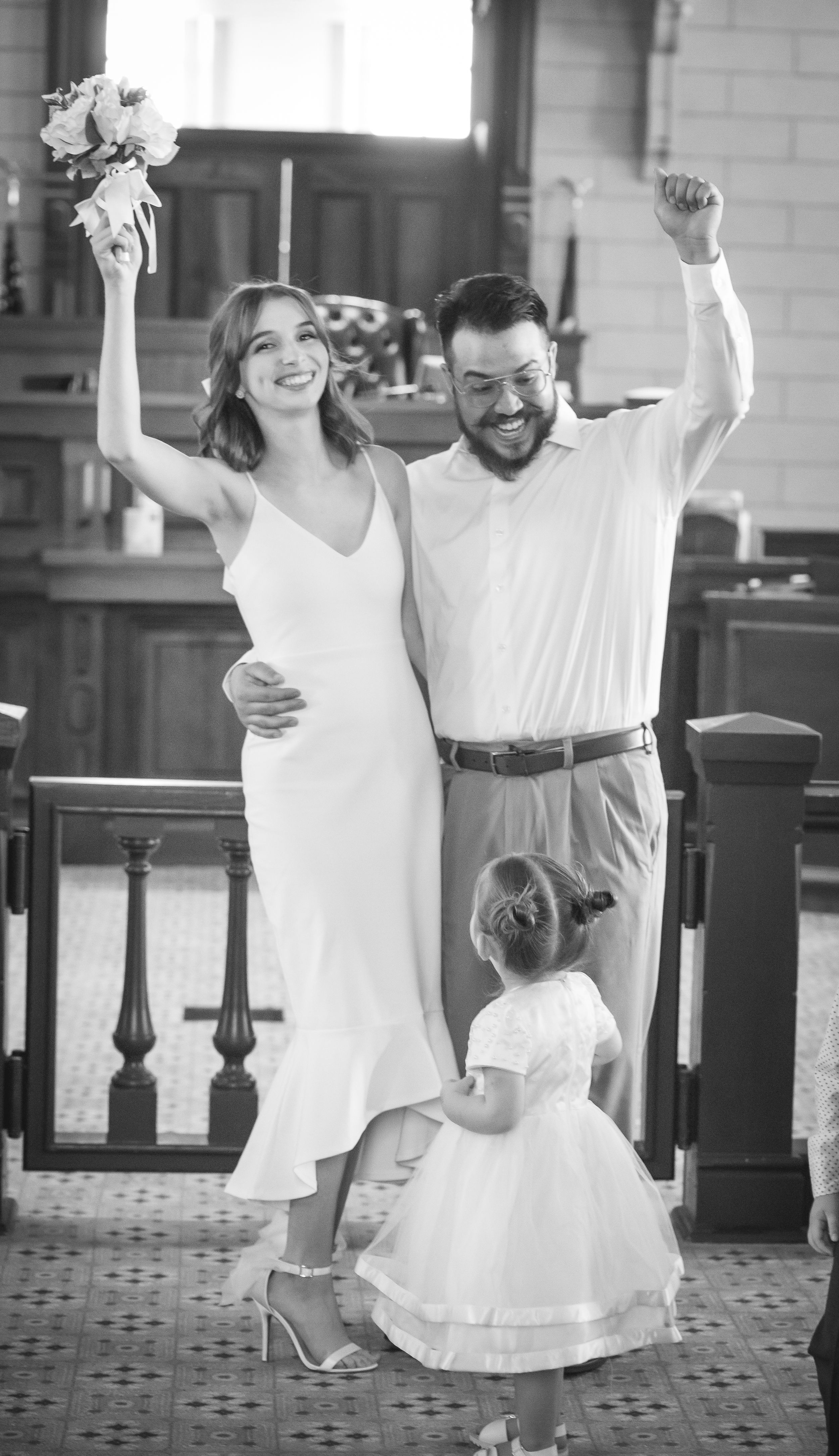 A joyous wedding celebration with a couple and small child in white attire raising their arms in celebration.