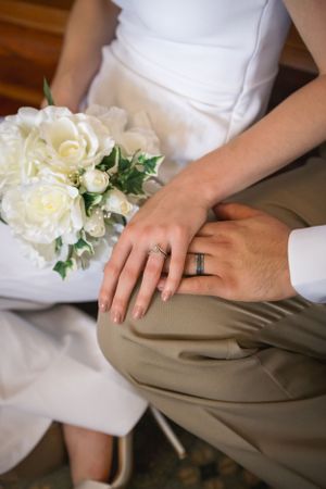Close-up of wedding rings and white rose bouquet with hands resting on khaki fabric.