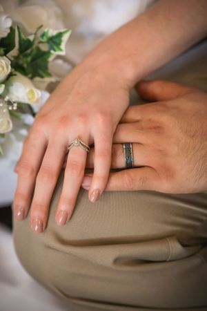 Close-up of newlyweds' hands showing their wedding rings with white roses in background.