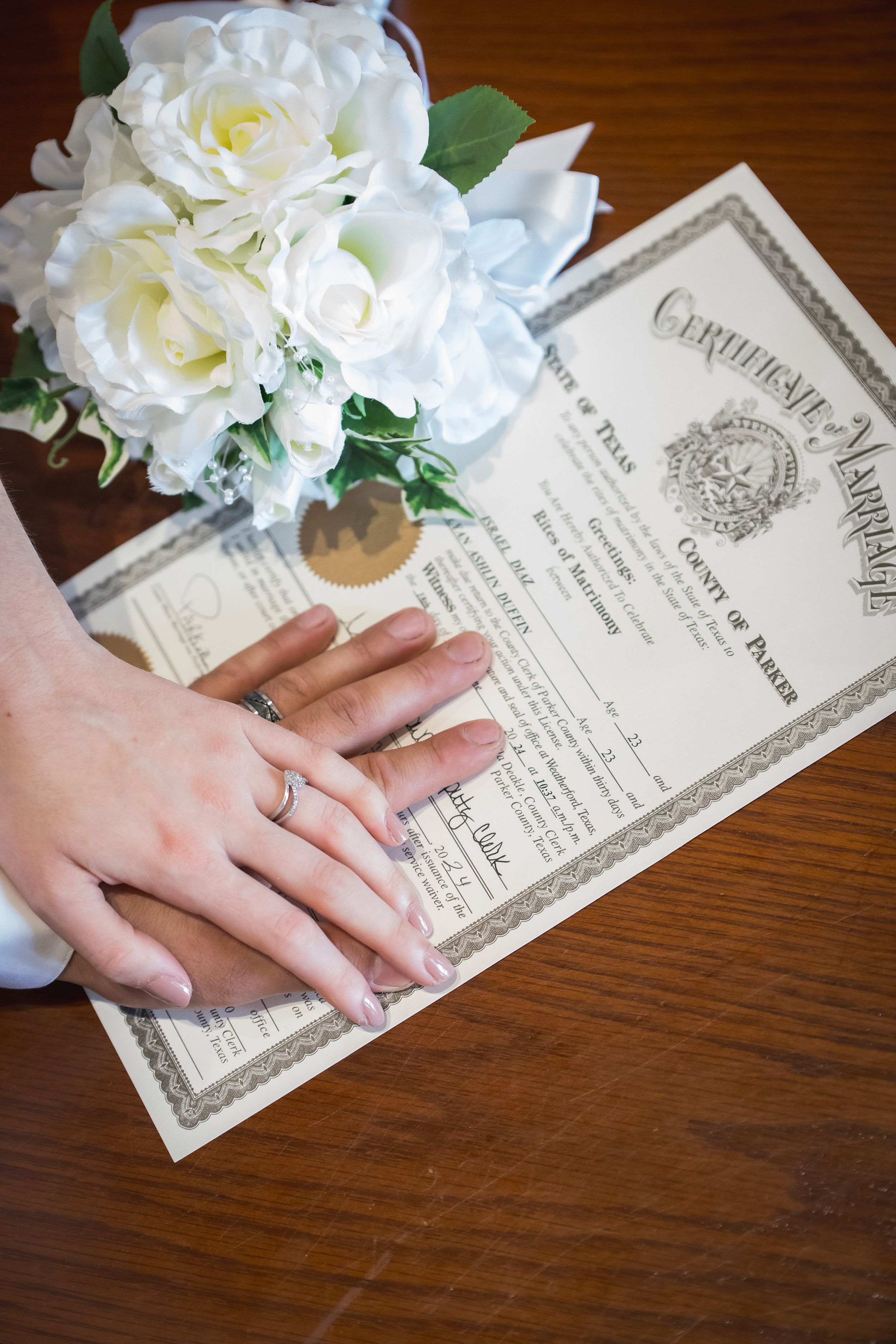 Wedding rings and white roses displayed with a marriage certificate on wooden surface.