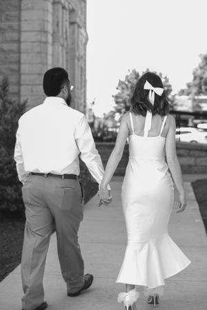 Black and white photo of newlyweds walking hand in hand along a sidewalk.