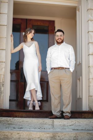 A couple poses on stone steps, with the woman wearing a white dress and the man in casual business attire.