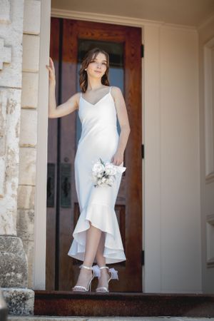 A person in a white fitted dress holds a small bouquet while posing on stone steps.