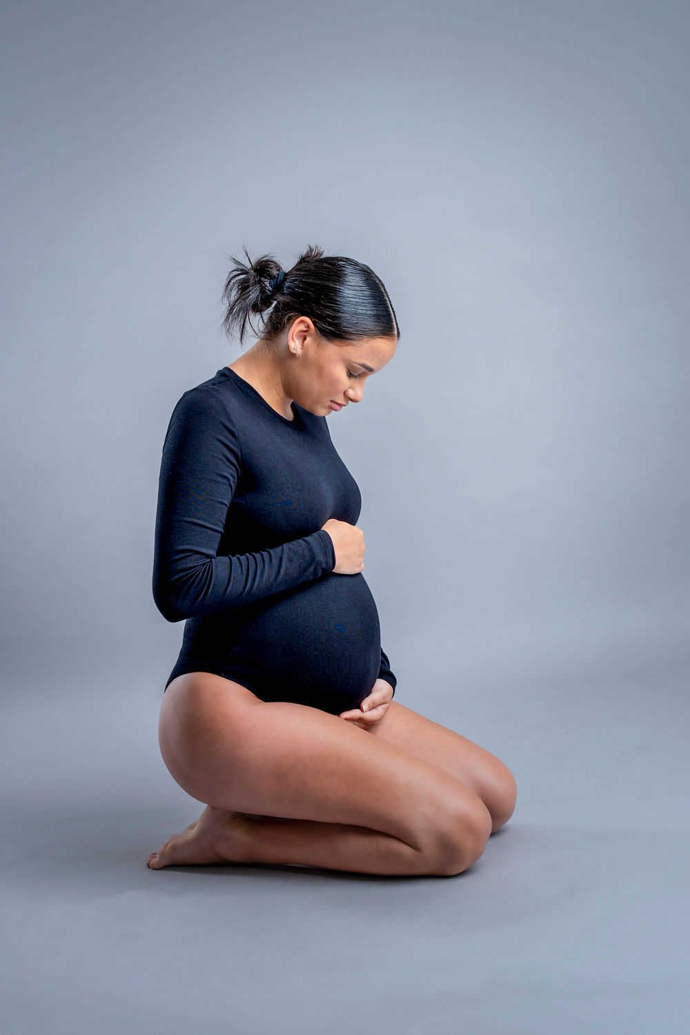 Une future maman vêtue d'un body bleu marine s'agenouille gracieusement sur un fond gris lors d'une séance photo de maternité.