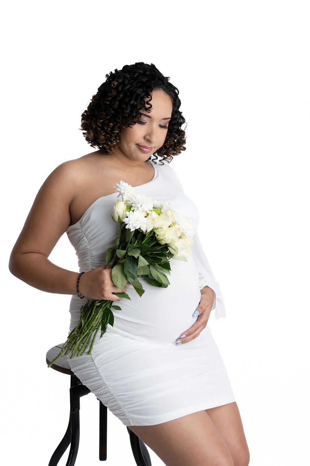 Élégante photo de maternité mettant en vedette une mini-robe blanche à une épaule et un bouquet crème sur fond blanc.