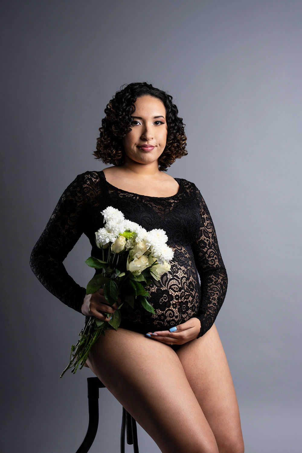 Un élégant portrait de maternité avec une silhouette en body en dentelle noire tenant des roses blanches sur un fond de studio gris.