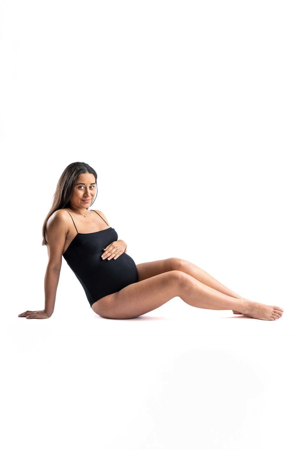 Une femme enceinte en body noir assise gracieusement sur un fond blanc tout en berçant son ventre rond.