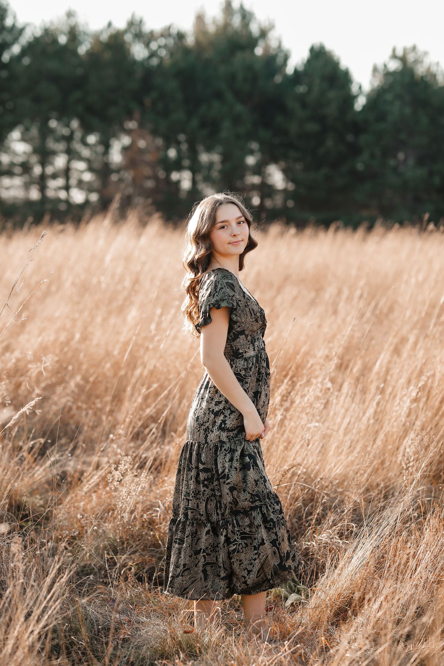 Young woman in floral maxi dress standing in golden wheat field with pine trees behind.