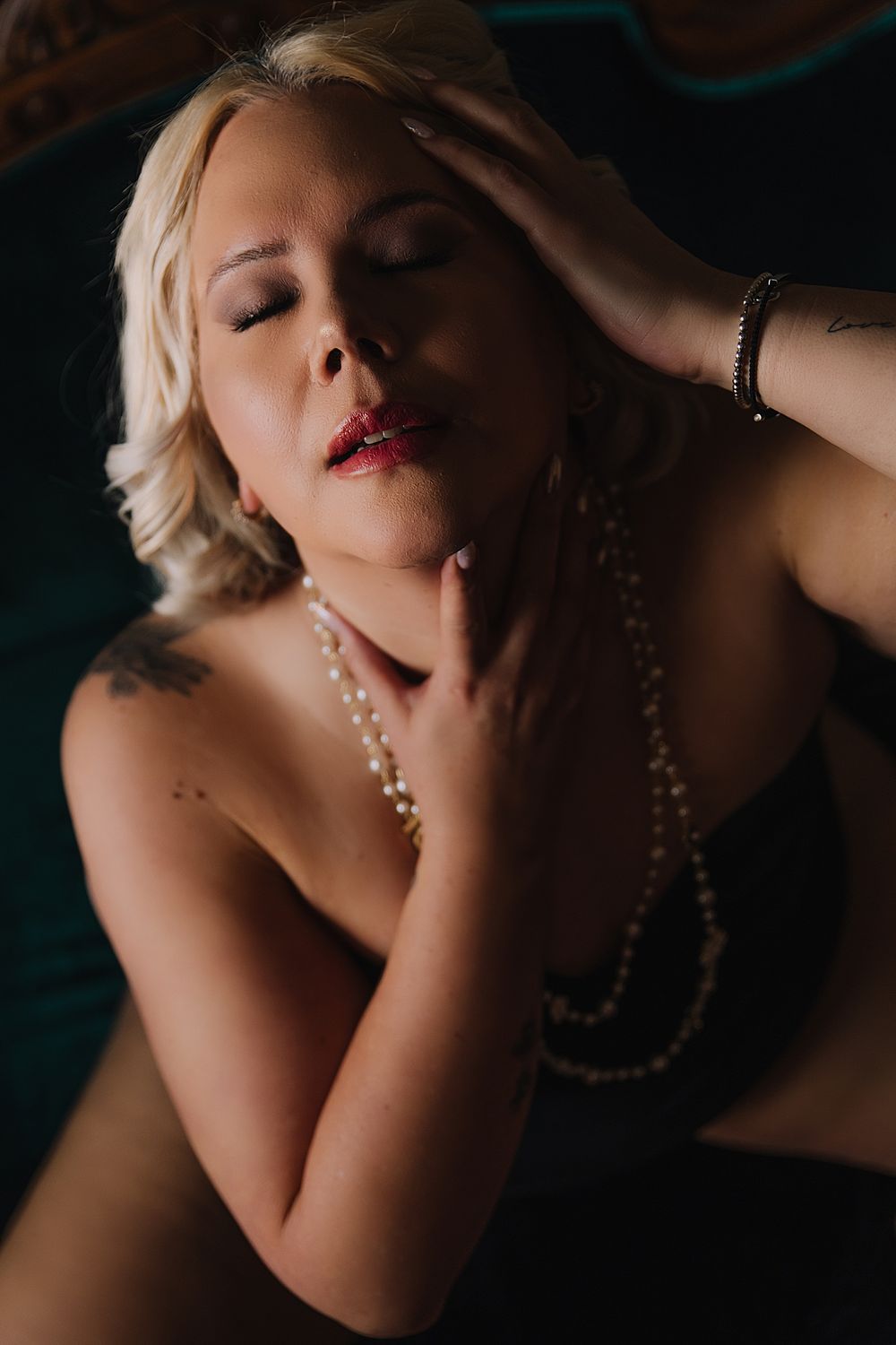 Sensual moody boudoir portrait featuring pearl necklace and red lipstick in dark dramatic lighting.