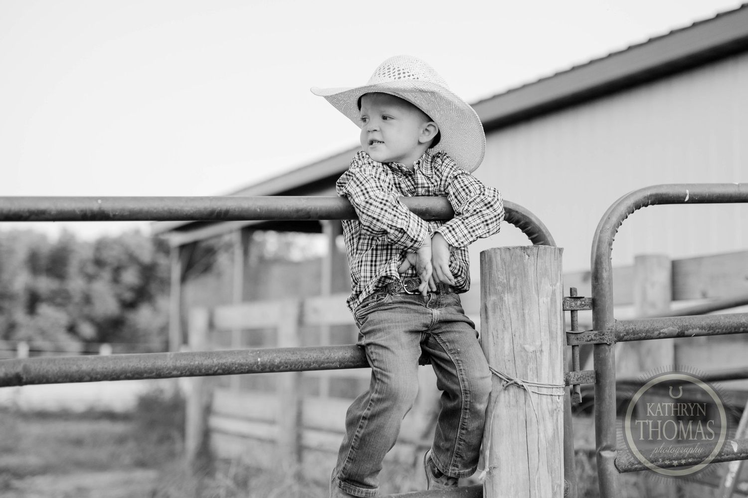 Black and white portrait of child in western wear sitting on wooden fence post.