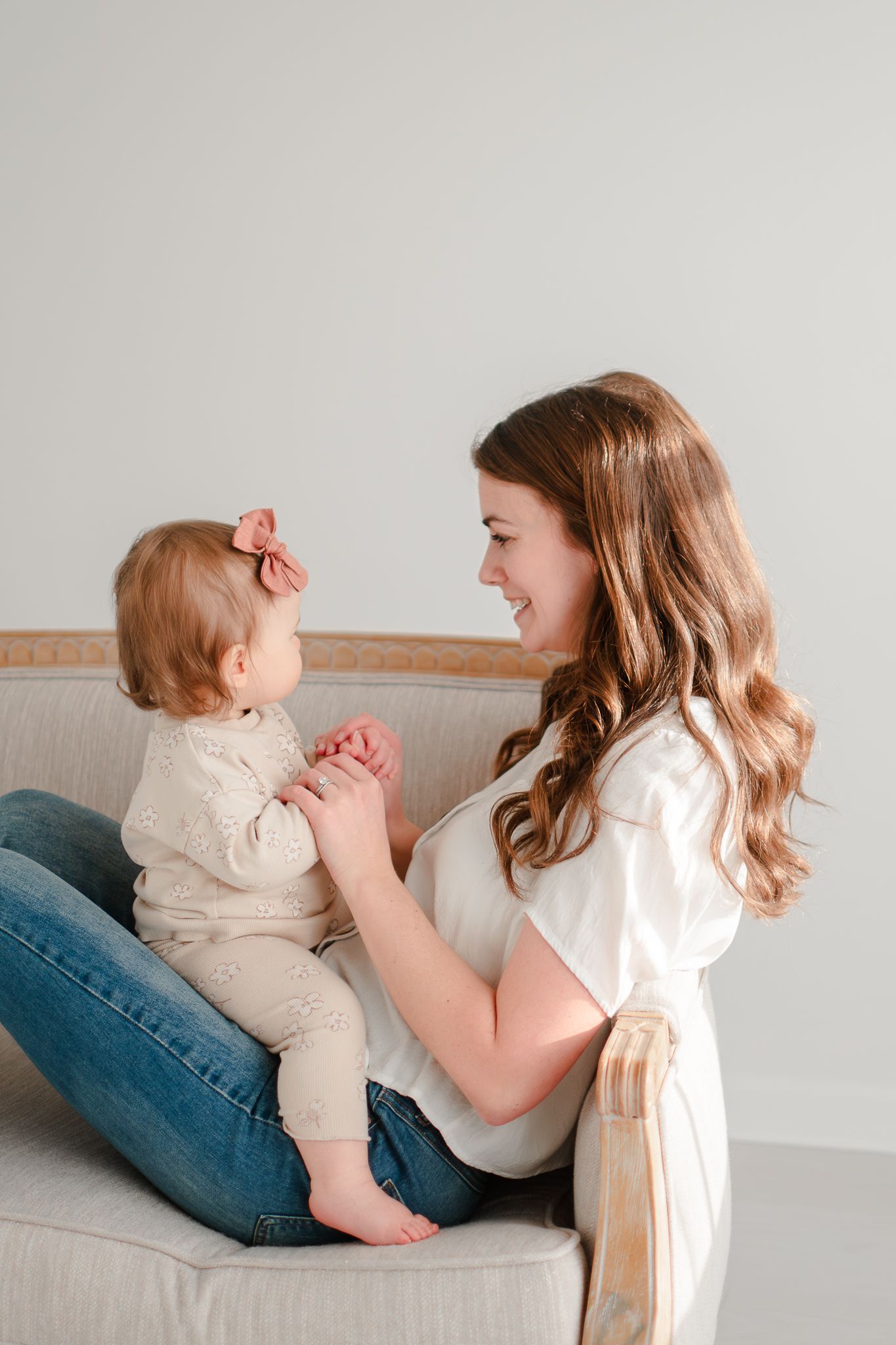 A tender moment shared on a gray couch as parent and toddler hold hands and smile at each other in a bright room.
