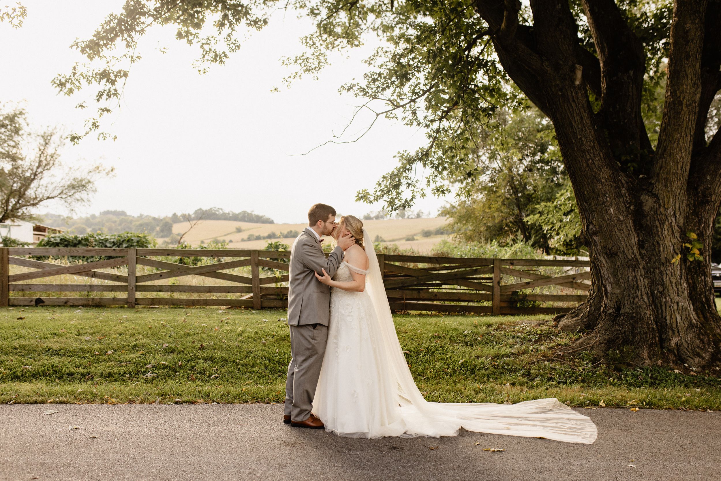 A couple shares an intimate moment at sunset by a rustic wooden fence under a large tree.