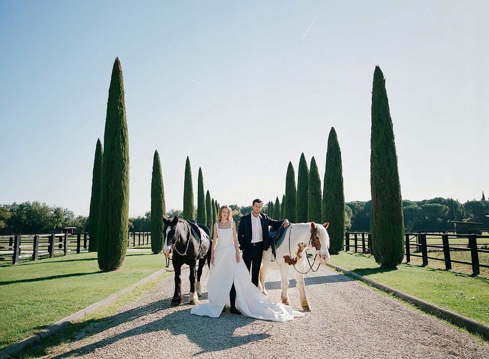 A wedding party walks down a cypress tree-lined path at an elegant countryside estate on a sunny day.