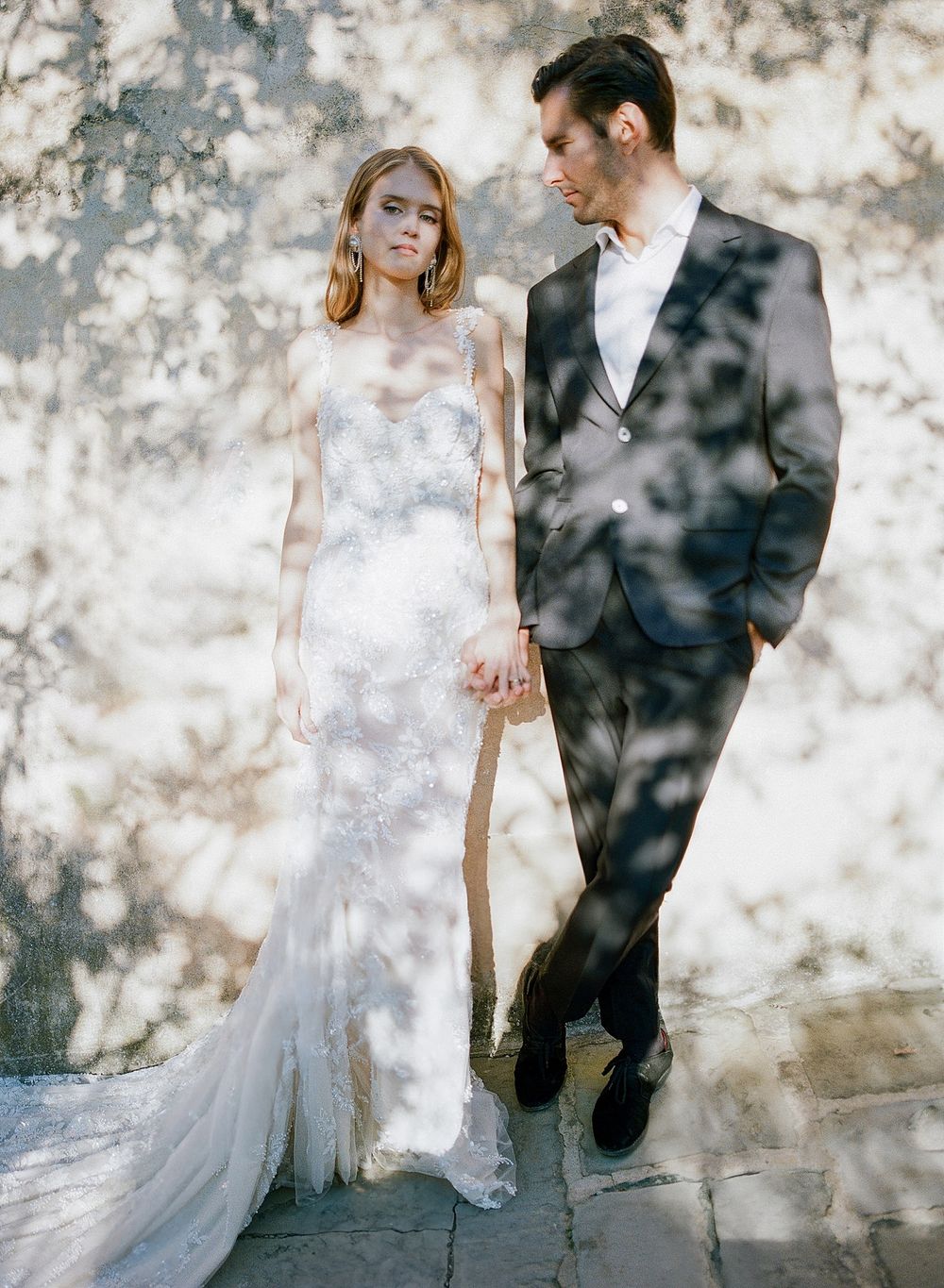 A romantic wedding photo with dappled tree shadows falling on a couple against a textured stone wall.