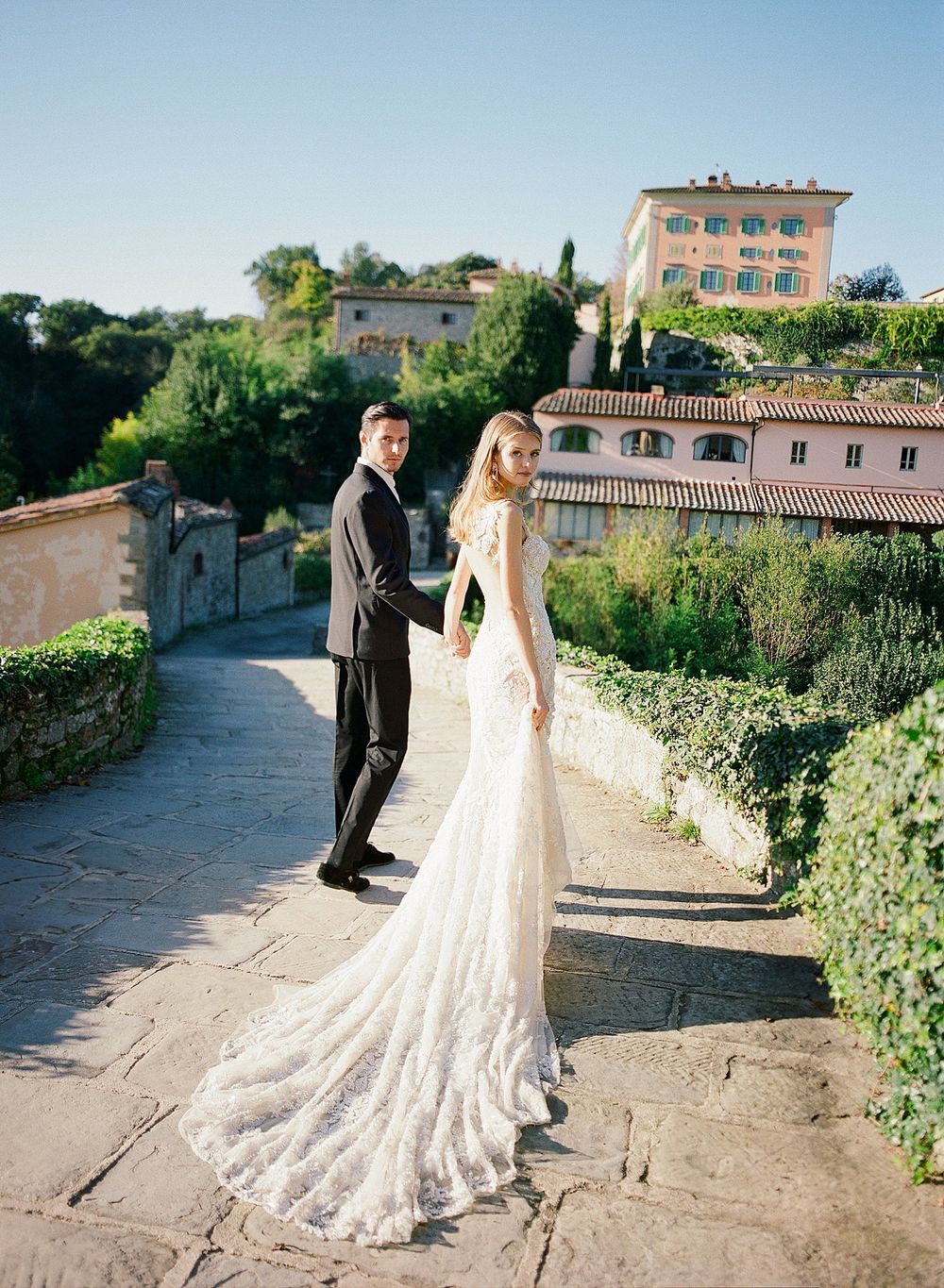 A wedding couple poses on a stone pathway with beautiful Italian villa architecture and gardens in the background.