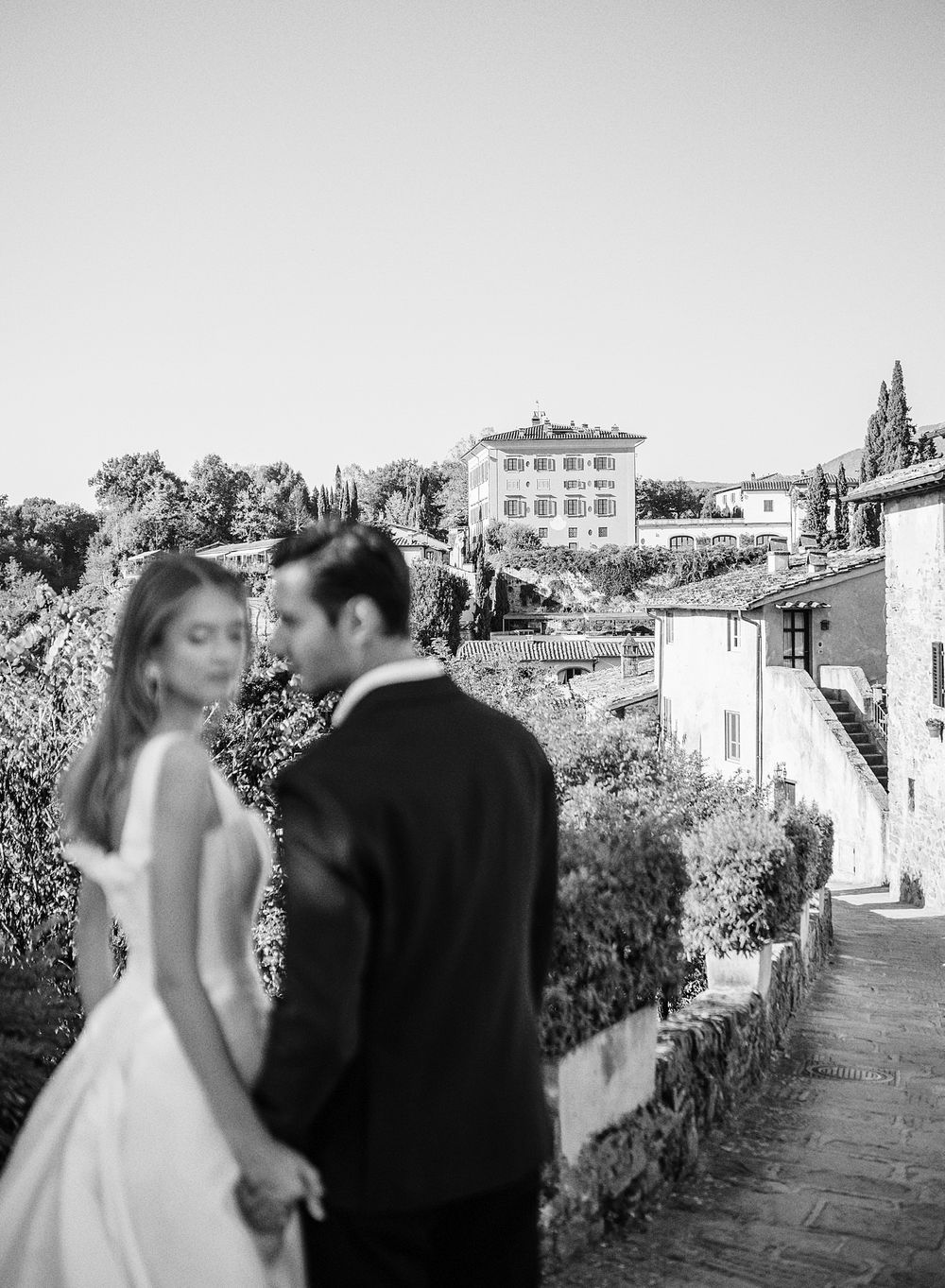 A couple in wedding attire share a romantic moment overlooking an Italian hillside villa in black and white.