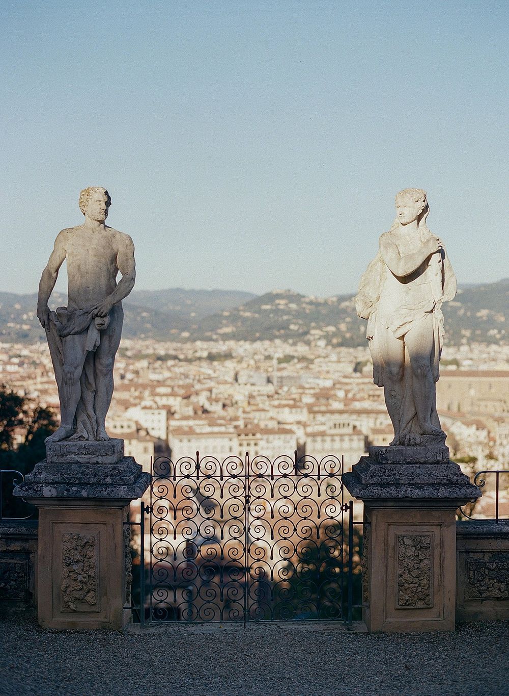 Two classical stone statues overlook a scenic view of Florence through an ornate iron gate on a terrace.
