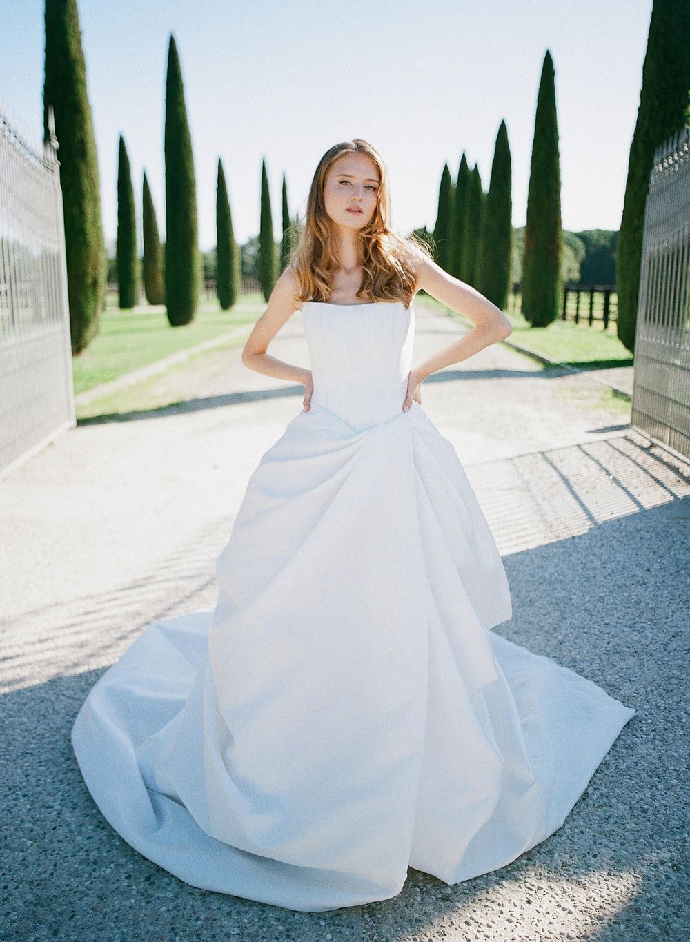 A stunning wedding gown with a voluminous skirt photographed in a sunlit garden path lined with Italian cypress trees.