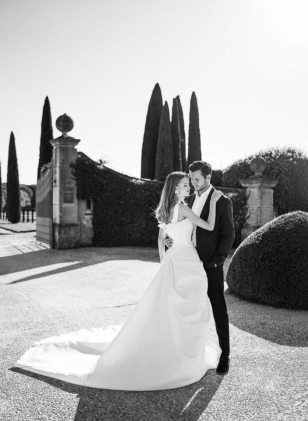 A romantic black and white wedding photo with tall cypress trees and stone architecture in an elegant garden setting.