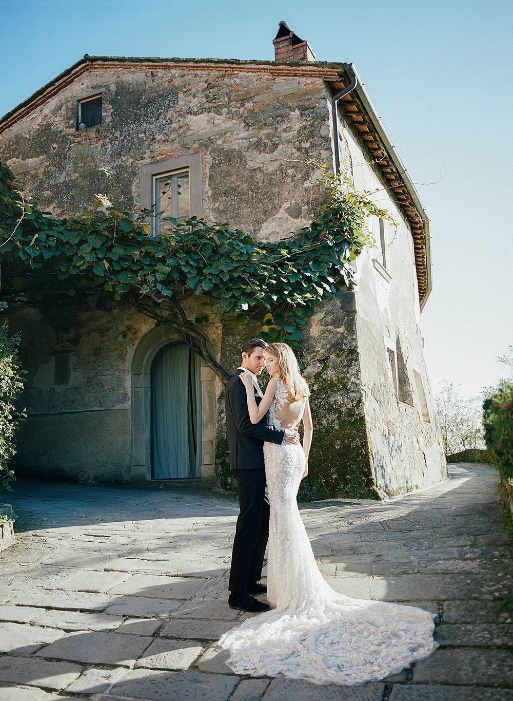 A romantic wedding couple embraces outside a rustic stone villa with climbing vines in an intimate countryside setting.