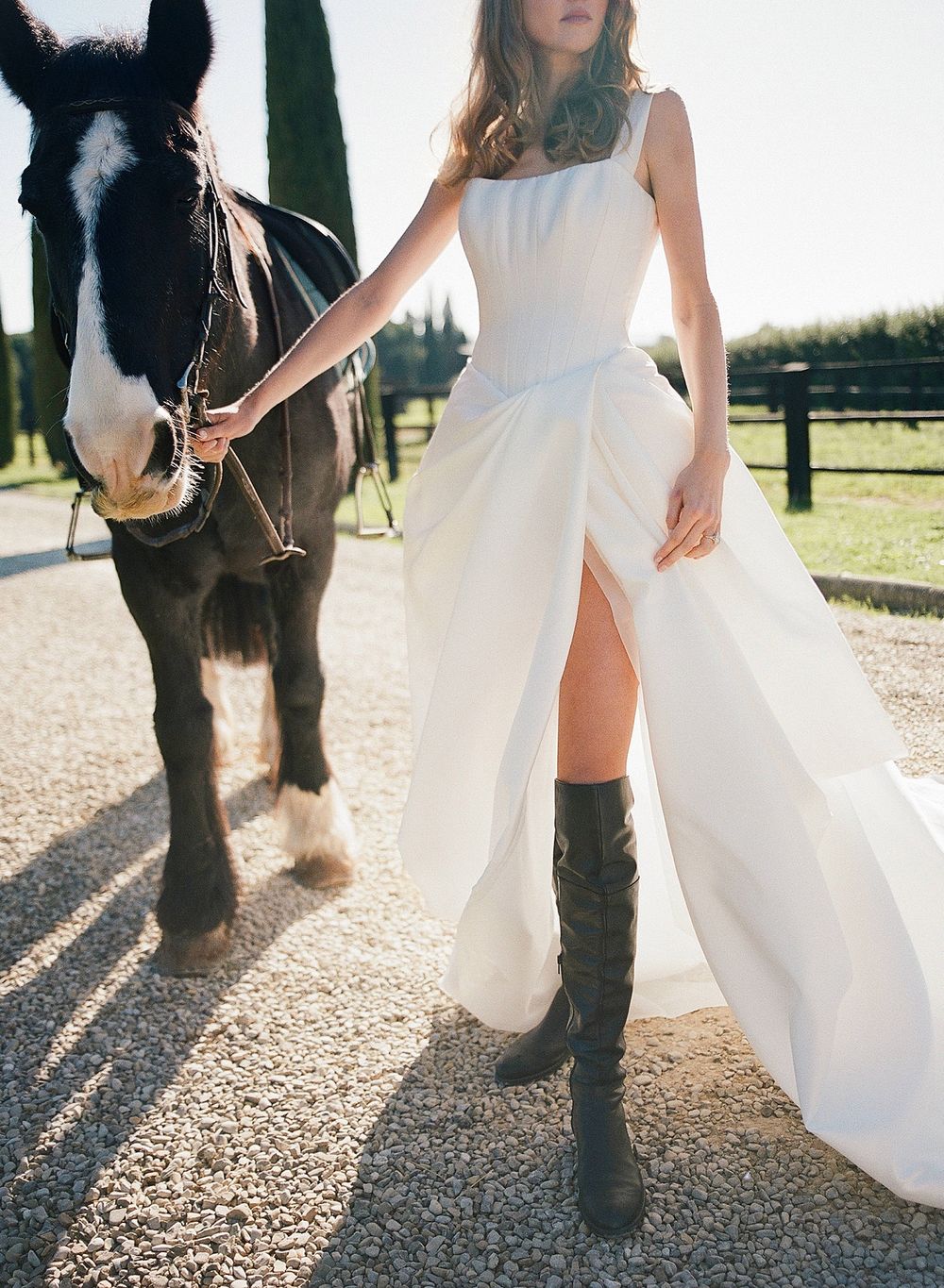 A white wedding dress and tall black riding boots paired with a black and white horse in a sunlit pasture setting.
