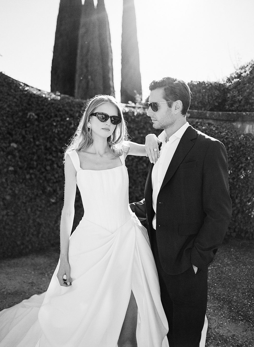 A stylish bride and groom wearing sunglasses pose together in a garden with architectural spires in the background.