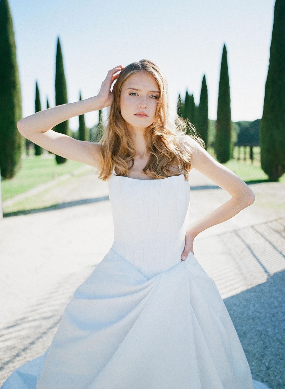 Elegant white wedding gown with fitted bodice and flowing skirt photographed in sunlight with cypress trees in background.