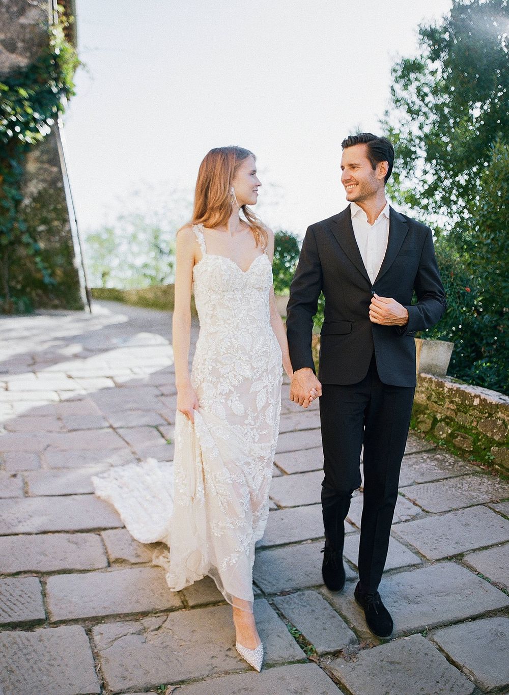 A couple in wedding attire walks together on a stone path while sharing a happy moment outdoors.