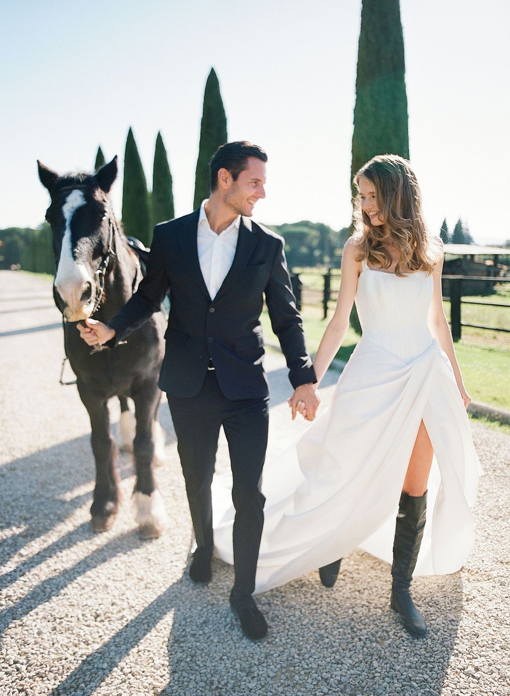 A romantic couple walks with a horse along a cypress-lined path in wedding attire, the bride wearing cowboy boots.